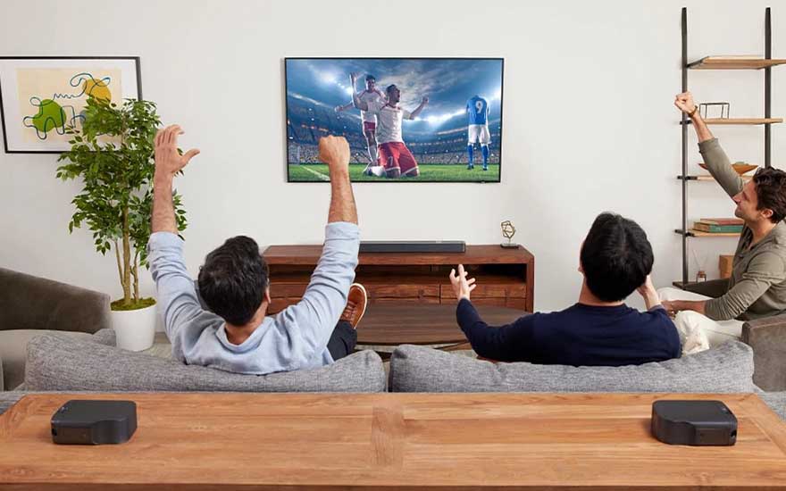 Drie mensen juichen op de bank tijdens voetbal op tv, met soundbar, draadloze speakers, plant en houten salontafel.