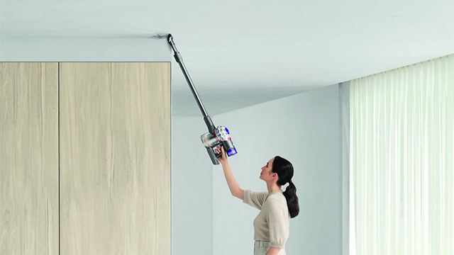 Een vrouw stofzuigt een plafond met een steelstofzuiger in een kamer met een kast en gordijnen.