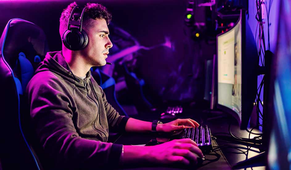 Gamer wearing headset sits focused at curved monitor, hands on keyboard and mouse, purple neon lighting fills the room.