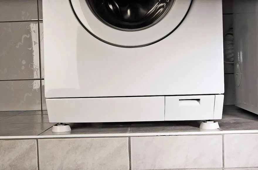 A white front-loading washing machine stands on a tiled platform. The lower compartment and adjustable feet are clearly visible. No brand is shown.