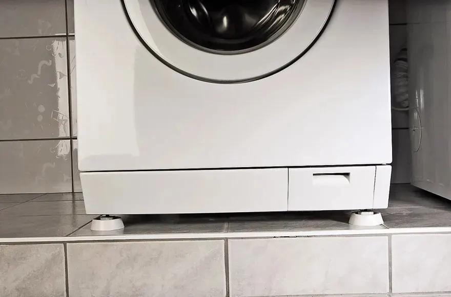 A white front-loading washing machine stands on a tiled platform. The lower compartment and adjustable feet are clearly visible. No brand is shown.