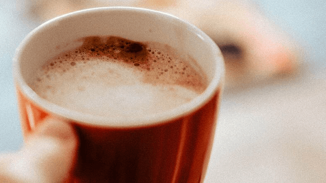 Een rode mok gevuld met een schuimende drank staat op tafel, vastgehouden door een hand. Op de achtergrond is een vaag object.