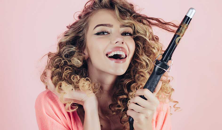Person with curly hair in pink outfit holding a curling iron against a pink background.