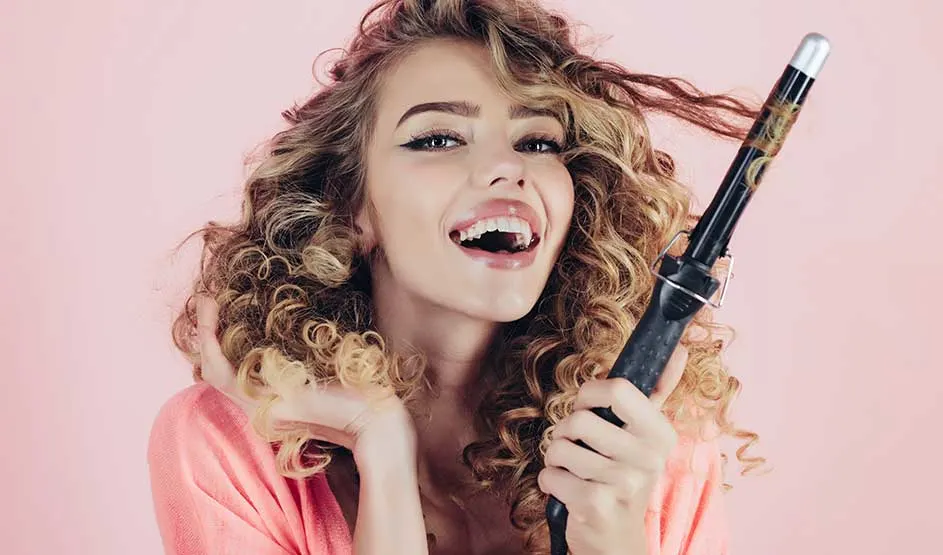Person with curly hair in pink outfit holding a curling iron against a pink background.
