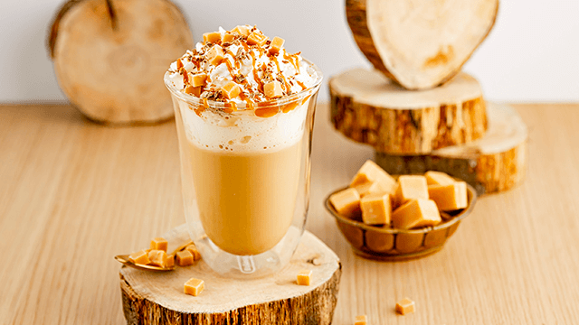 Een glas karamellatte met slagroom en karamelblokjes op een houten onderzetter, met karamelblokjes in een kom.