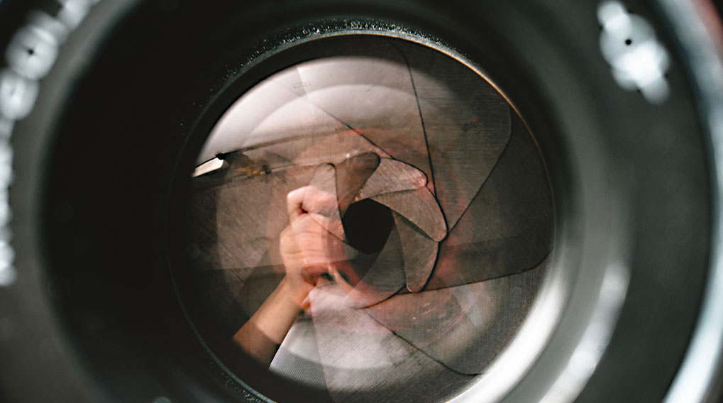 Een close-up van een lens met een diafragma en een reflectie van een hand en een witte achtergrond.