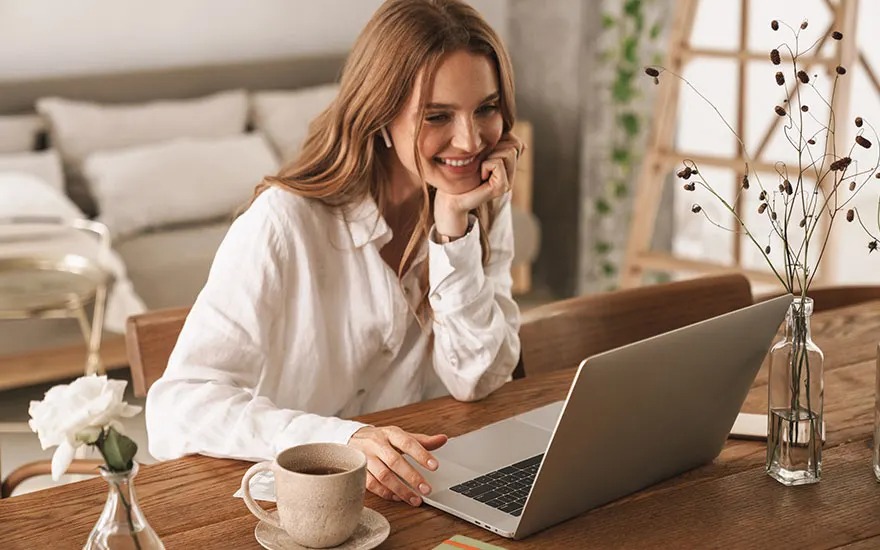 Woman smiles at laptop on wooden table, with coffee cup and flowers in vases, in a warm, cozy living space.