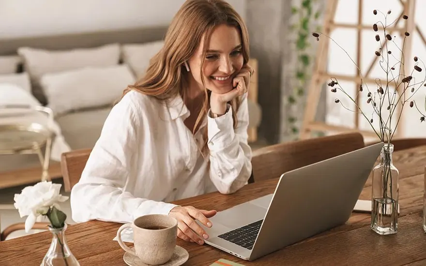 Woman smiles at laptop on wooden table, with coffee cup and flowers in vases, in a warm, cozy living space.