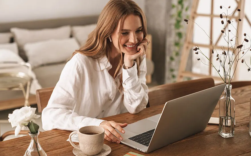 Glimlachende vrouw in wit shirt werkt aan laptop, koffiekop en bloemen op houten tafel, moderne huiskamer achtergrond