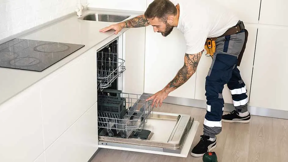 Een man met gereedschap opent een vaatwasser in een witte keuken met een kookplaat en een gootsteen.