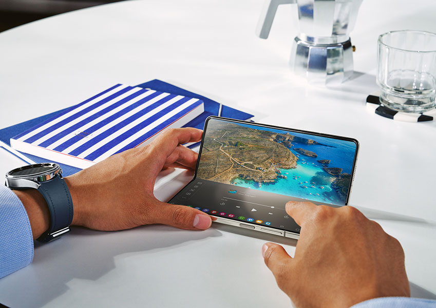 Handen bedienen een opvouwbare tablet met een kustlandschap op het scherm, naast notitieboekjes, een horloge en een espressomaker.