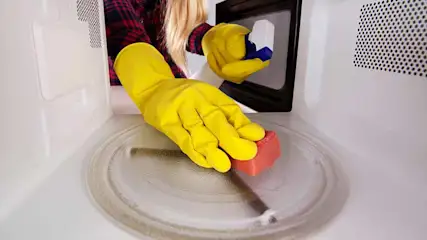 Een hand met een gele rubberen handschoen maakt de binnenkant van een witte magnetron schoon met een roze spons.