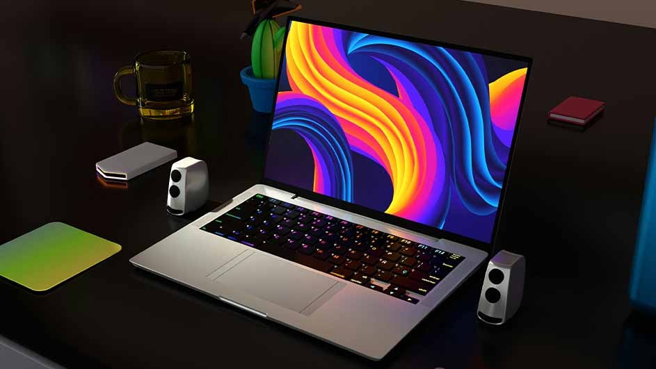 Laptop displays a colorful screen, flanked by speakers. A mug, cactus, book, and mousepad sit on a dark desk.