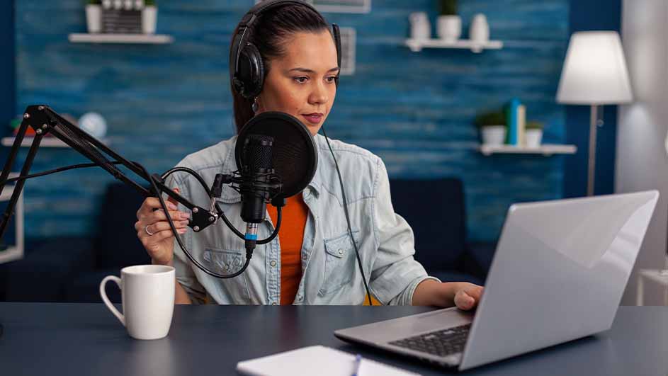 Een vrouw met een koptelefoon zit aan een tafel met een microfoon, laptop, notitieblok en een witte mok. De achtergrond is blauw.