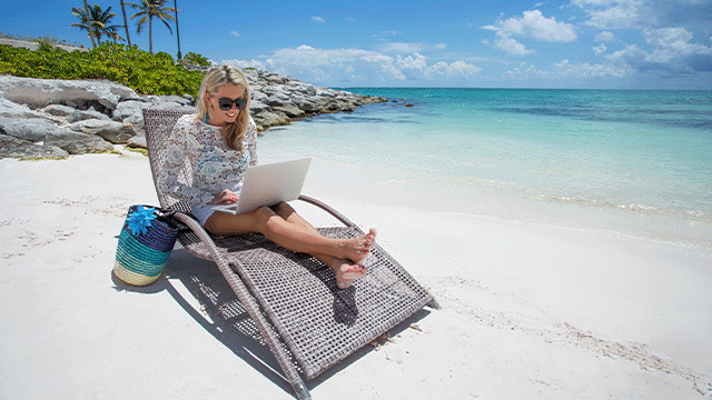 Vrouw zit op een stoel op het strand met een laptop. Een tas staat naast haar. De zee is blauw en er zijn palmbomen.