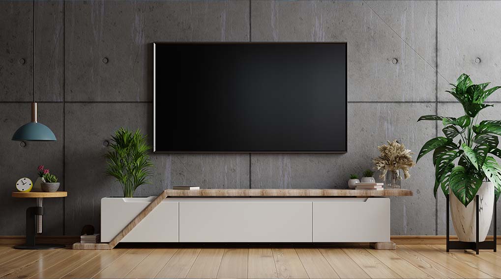 Betonnen muur met tv erboven. Een wit dressoir met planten, decoratie en een kleine tafel links. Houten vloer.