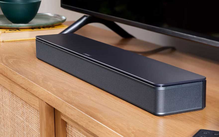 Close-up van zwarte Bose soundbar op houten tv-meubel, naast schermvoet, kom, boek en afstandsbediening.
