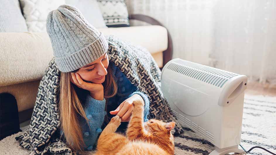 Een vrouw met een grijze muts en deken speelt met een oranje kat bij een witte kachel op een tapijt.