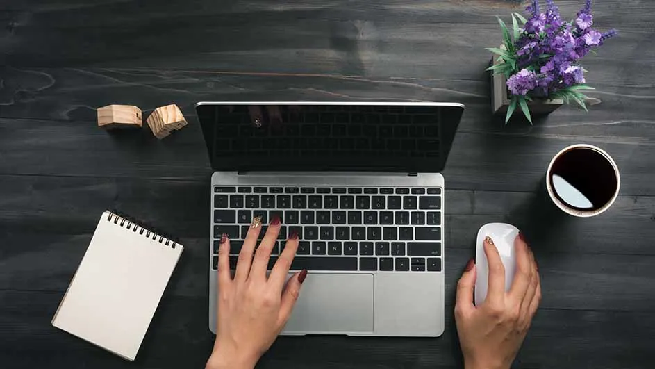 Een bureau met een laptop, notitieboek, houten blokken, een kop koffie, een bloempot en handen die een muis en toetsenbord bedienen.