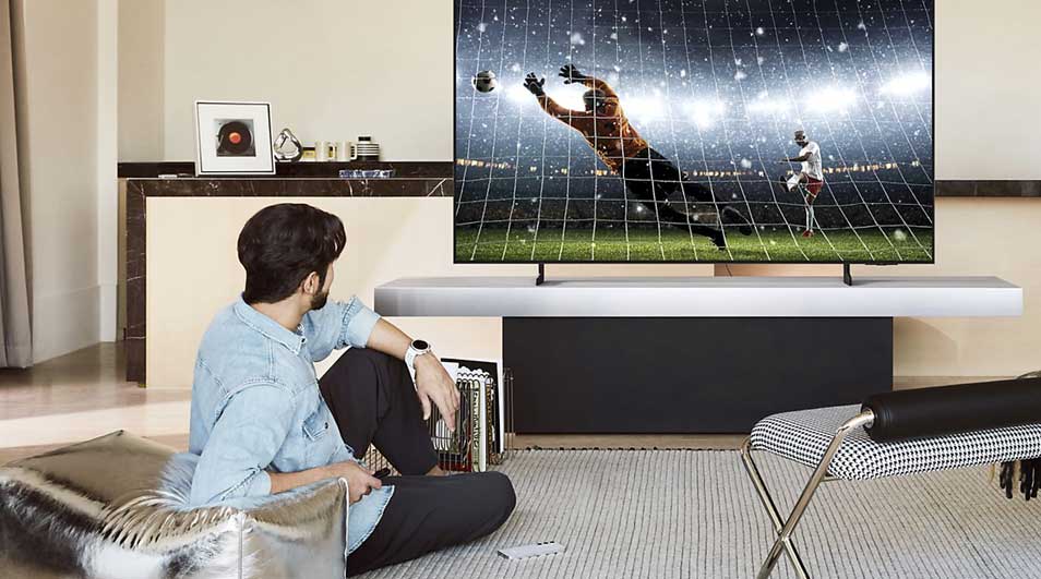 A man sits on the floor watching a large wall-mounted television displaying a football match. Modern interior with wooden furniture, grey curtains, neutral tones. Minimalist, stylish living room setup.
