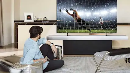 Een man zit op de vloer en kijkt naar een groot wandmontage-tv met voetbalwedstrijd. Modern interieur met houten meubels, grijze gordijnen, licht. Minimalistische, stijlvolle woonkamer setup.