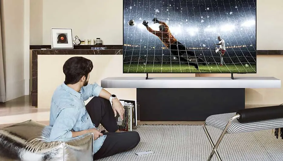 Een man zit op de vloer en kijkt naar een groot wandmontage-tv met voetbalwedstrijd. Modern interieur met houten meubels, grijze gordijnen, licht. Minimalistische, stijlvolle woonkamer setup.