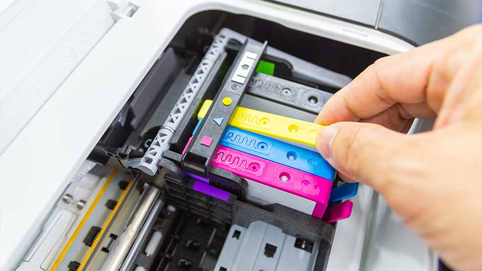 Een hand plaatst een gele inktcartridge in een printer met roze, blauwe en zwarte cartridges.