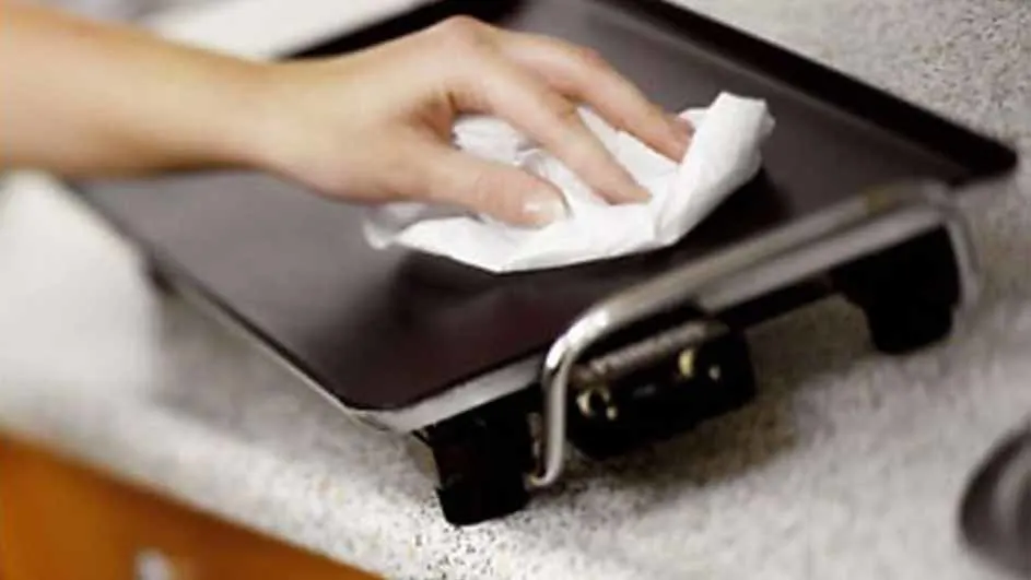Een hand veegt een platte, zwarte grill schoon met een wit papieren doekje. De grill staat op een aanrecht met spikkels.