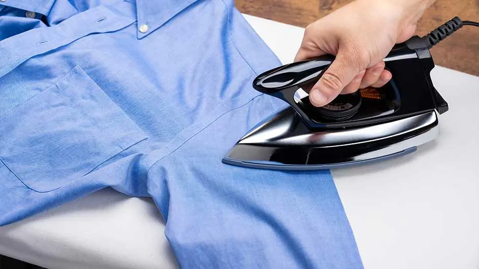 Een hand strijkt een blauw overhemd met een zwart strijkijzer op een wit oppervlak.