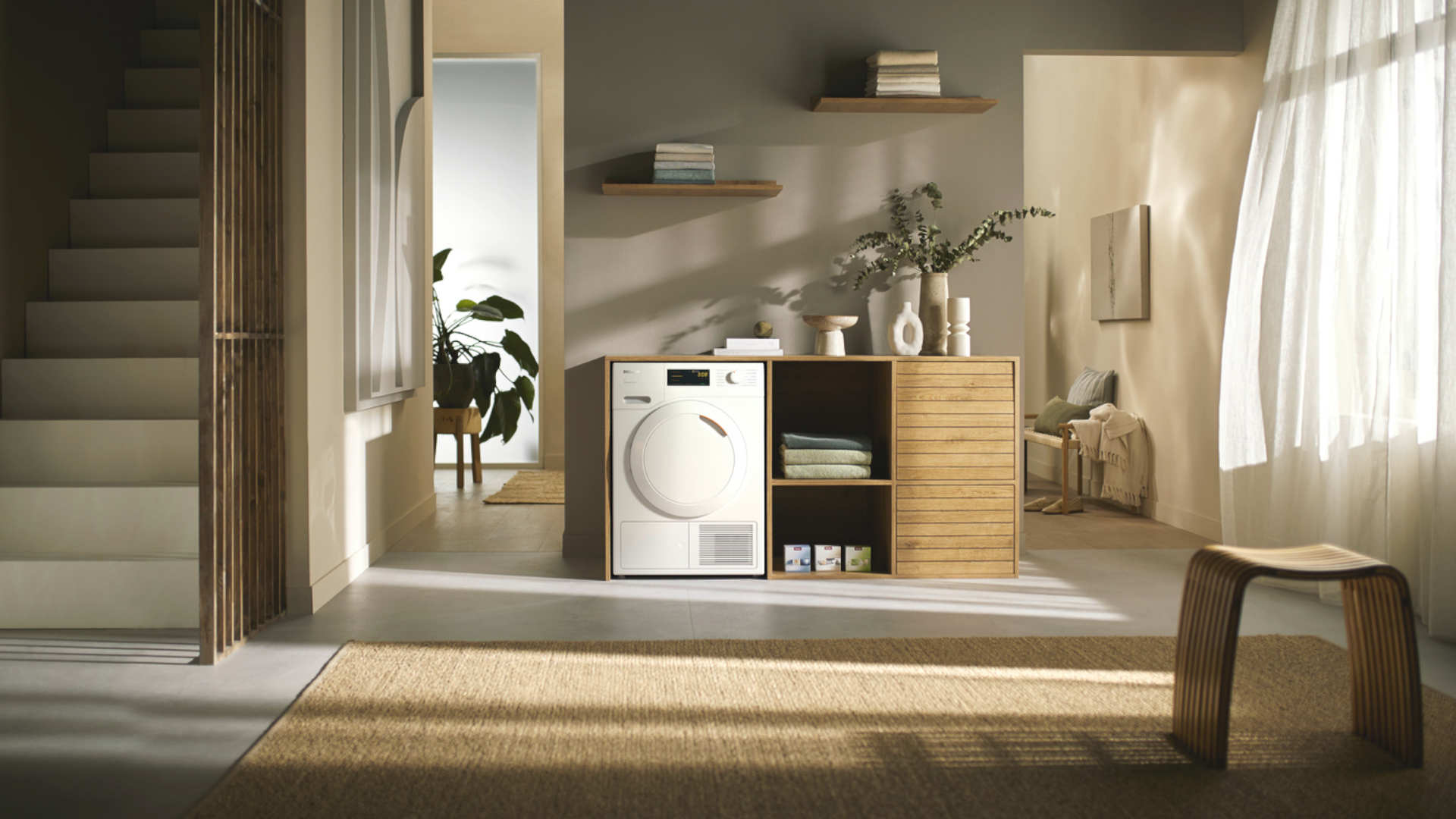 Woonkamer met houten trap, kast met wasmachine, decoratie en stoel, verlicht door een raam met gordijnen.