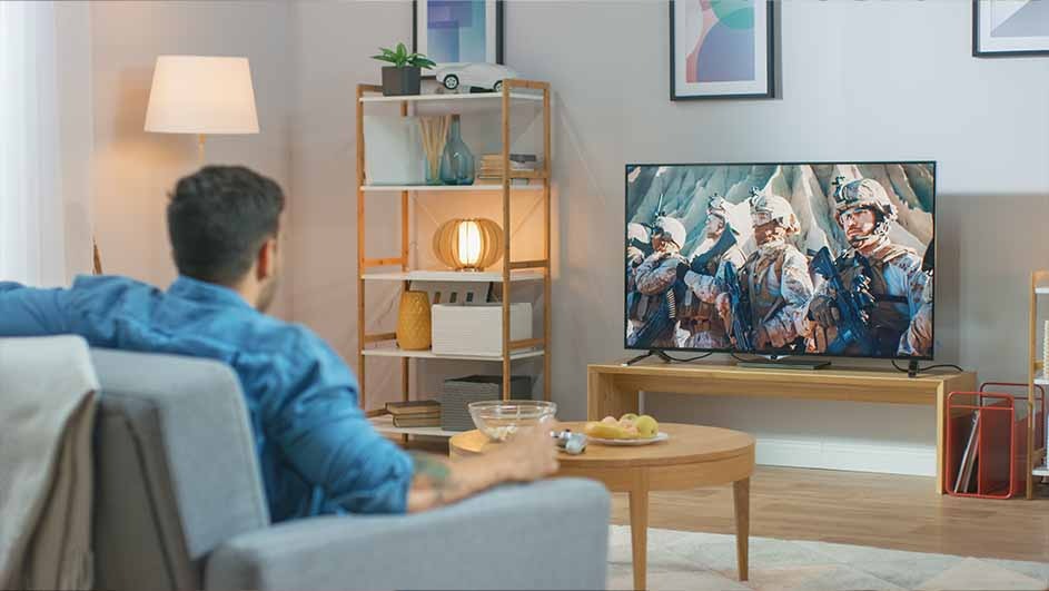A person sits on a gray couch watching a television showing soldiers. A table with snacks and a bookshelf are nearby.