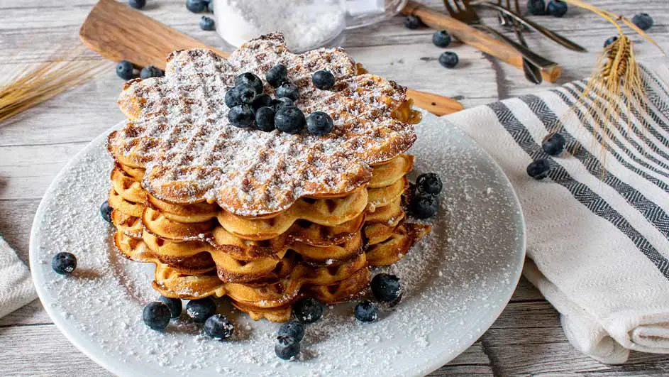 Een stapel bloemvormige wafels, bestrooid met poedersuiker en blauwe bessen, op een wit bord.