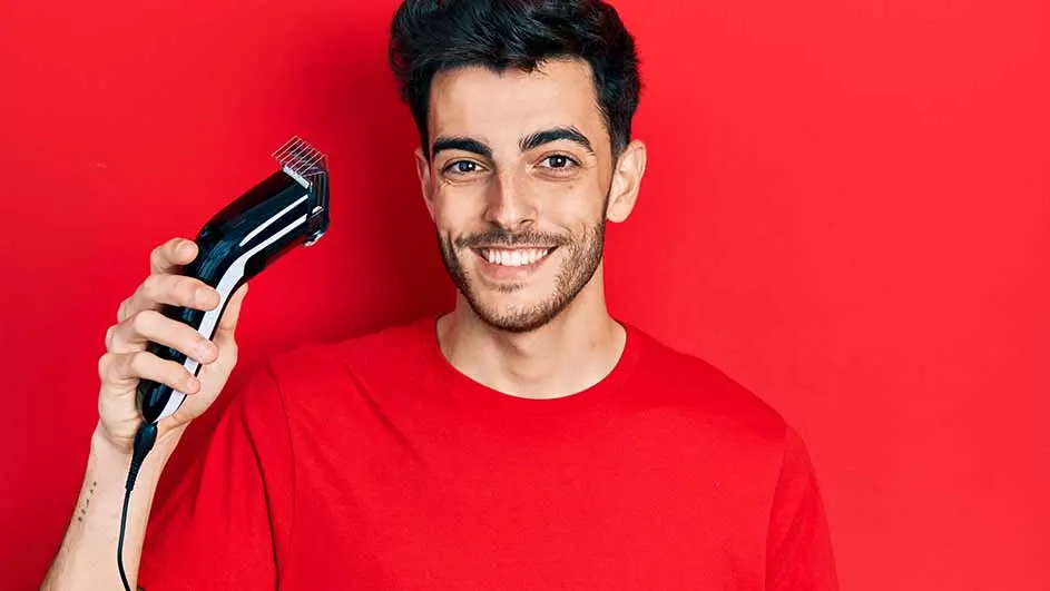 Een man met een rode trui glimlacht en houdt een zwarte tondeuse met een snoer vast tegen een rode achtergrond.