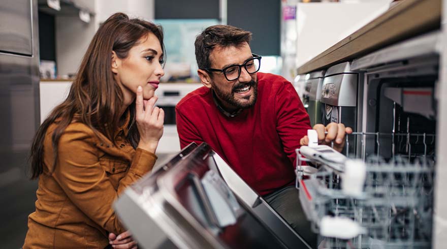 Een man in een rode trui en een vrouw in een bruine blouse bekijken een zilverkleurige vaatwasser in een winkel.