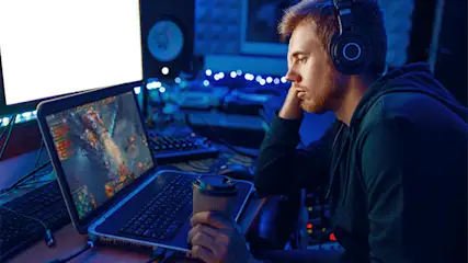 Een man met een koptelefoon kijkt naar een laptop met een spel. Hij heeft een beker in zijn hand en zit aan een bureau.