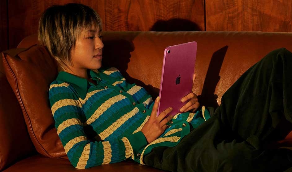 Person relaxing on a brown leather sofa, holding a purple tablet upright, lit by warm light with wooden wall in background.