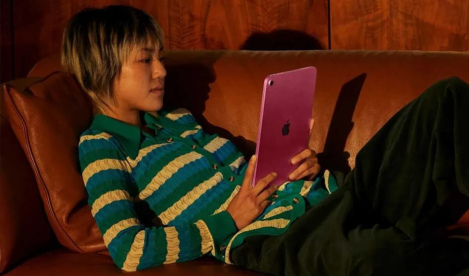 Person relaxing on a brown leather sofa, holding a purple tablet upright, lit by warm light with wooden wall in background.