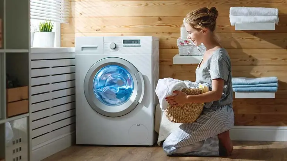 Een vrouw in grijze kleding knielt bij een moderne witte wasmachine met blauw verlichte trommel. Ze houdt een rieten mand met wasgoed vast in een wasruimte met houten wanden, opgerolde handdoeken en een plantje.