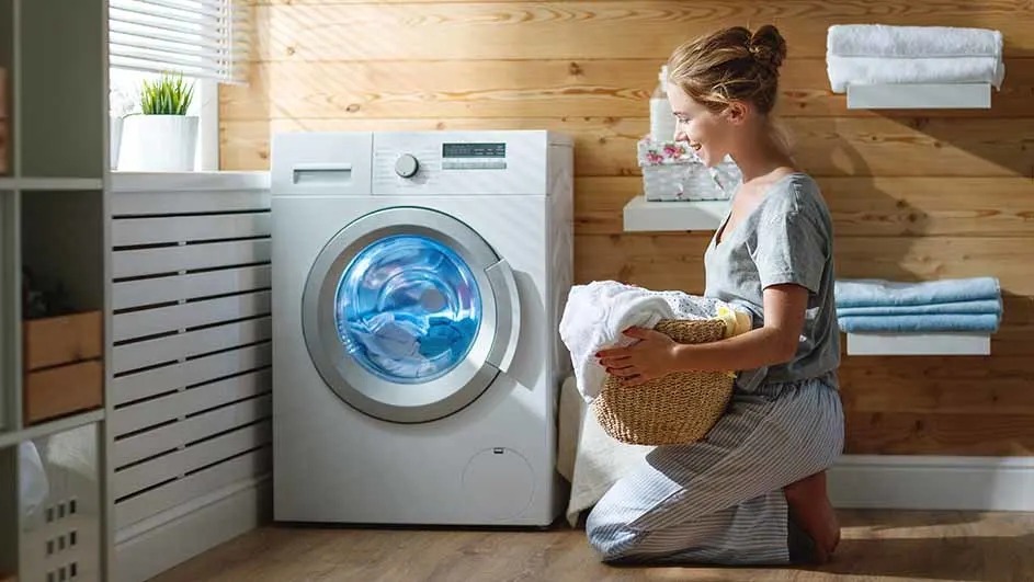 A woman kneels in a cosy laundry room, holding a wicker basket while loading clothes into a modern white washing machine. No brand is visible.