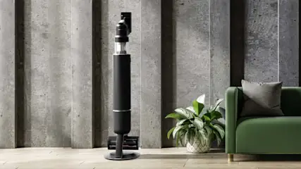 Een zwarte draadloze stofzuiger staat naast een groene bank en een plant tegen een betonnen muur.