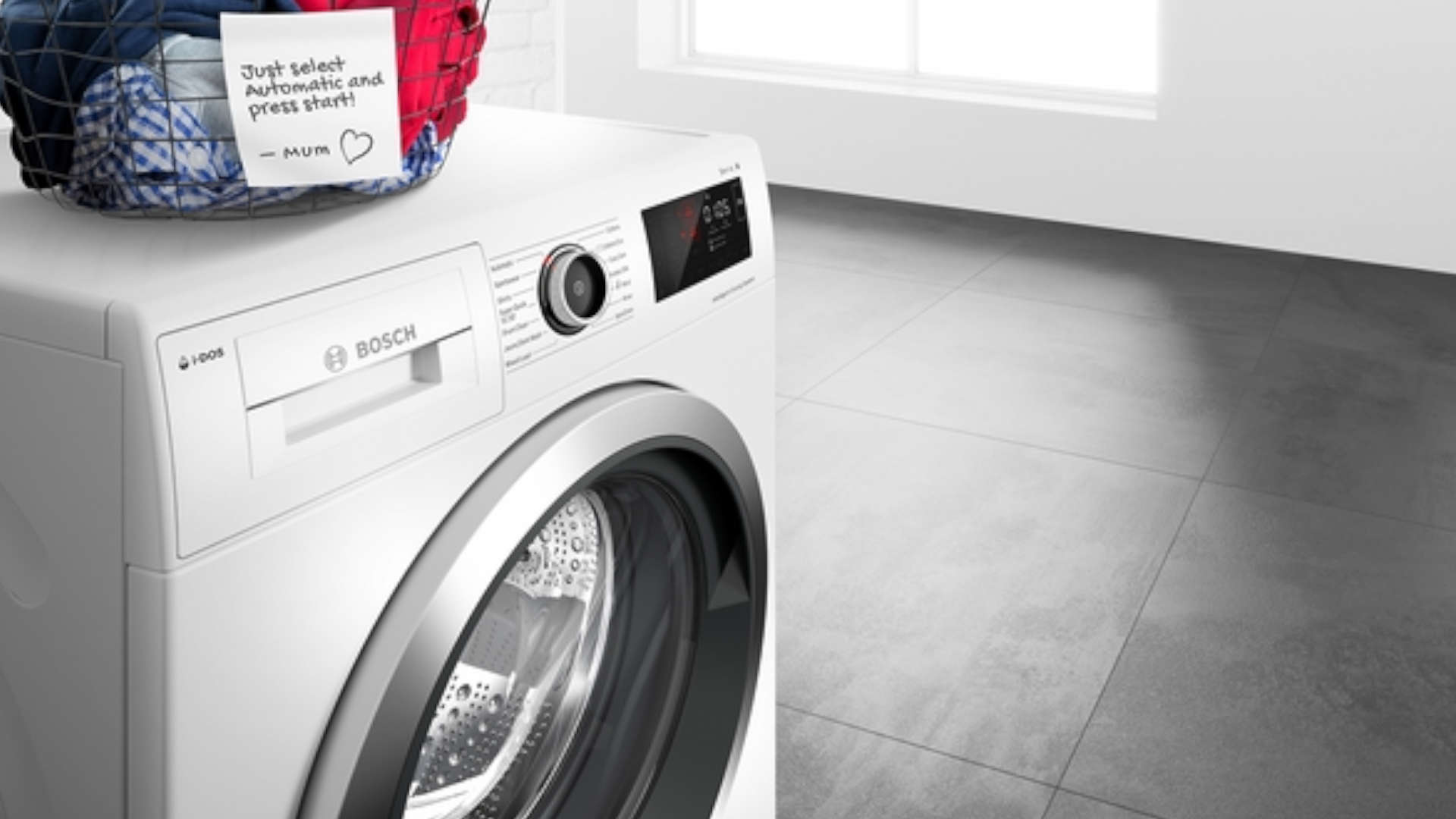 Bosch wasmachine met wasmand erop in een kamer met grijze tegels. Een briefje aan de wasmand met instructies.