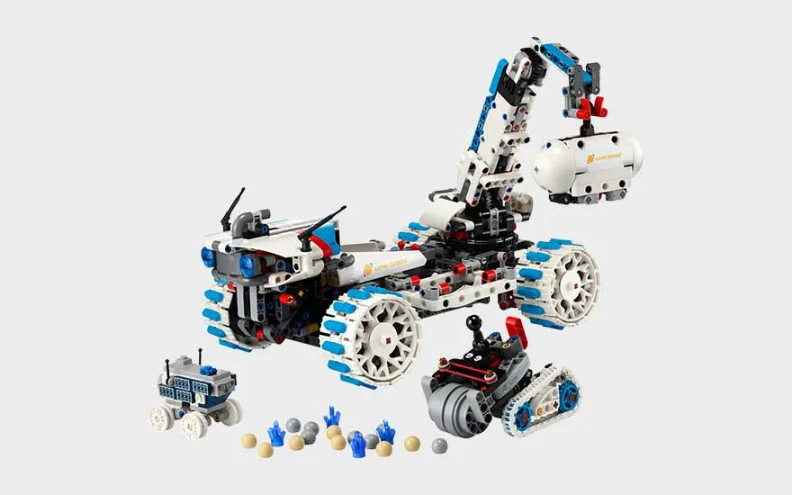 Witte LEGO ruimterover met kraanarm, grote wielen, kleine voertuigen en accessoires voor een maanmissie op grijze achtergrond.