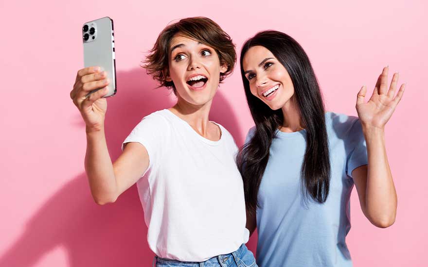 Two young women happily take a selfie in front of a pink background; one holds the phone and the other smiles and waves at the camera.
