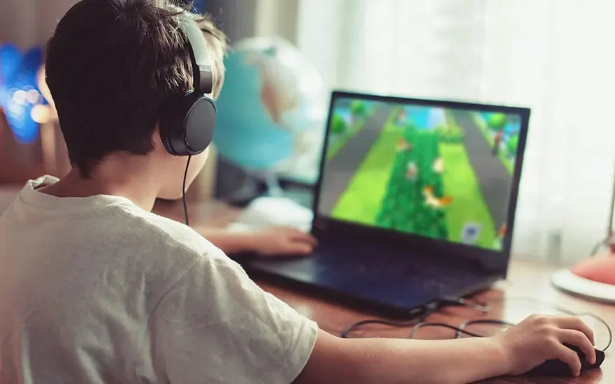 Child wearing headphones plays a colorful video game on a laptop at a desk, hand on mouse, globe and lamp in the background.