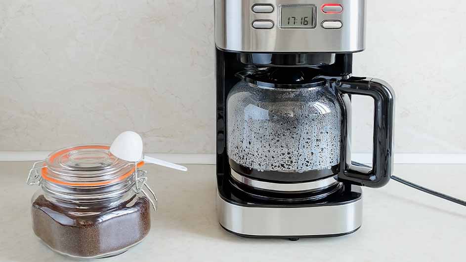 Filter coffee machine on a kitchen counter next to a jar of ground coffee with a measuring spoon.
