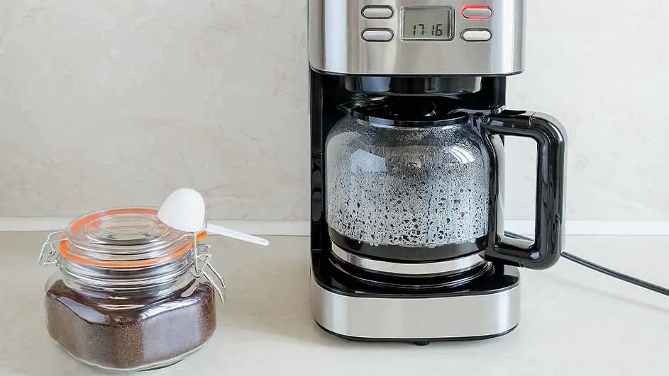 Filter coffee machine on a kitchen counter next to a jar of ground coffee with a measuring spoon.