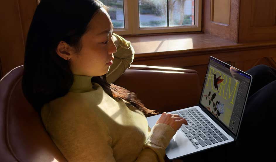 Student lounges in a leather chair by the window, sunlight on her face as she calmly works on a colourful graphic design on her laptop.