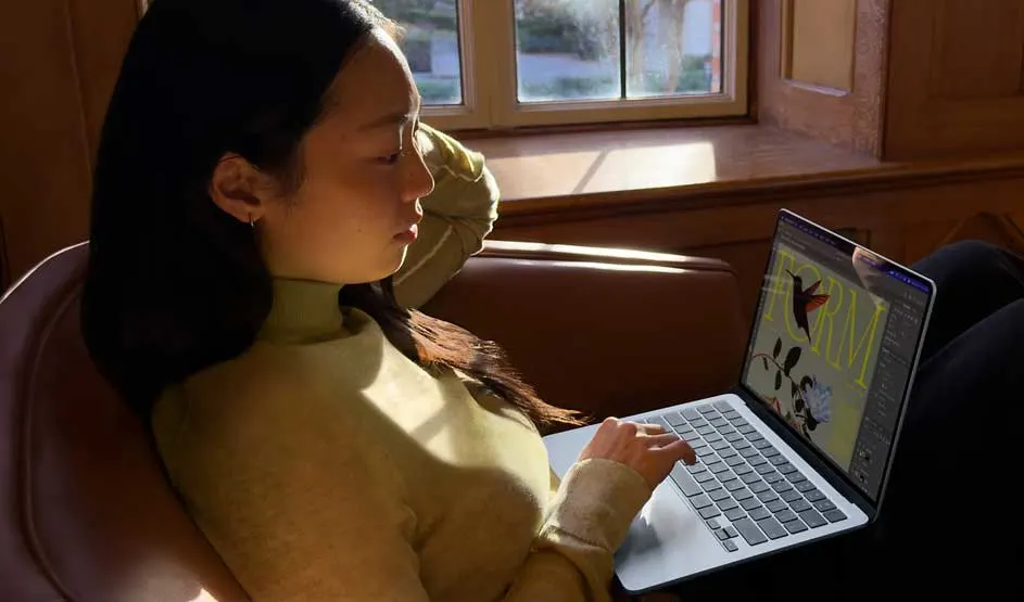 Student lounges in a leather chair by the window, sunlight on her face as she calmly works on a colourful graphic design on her laptop.