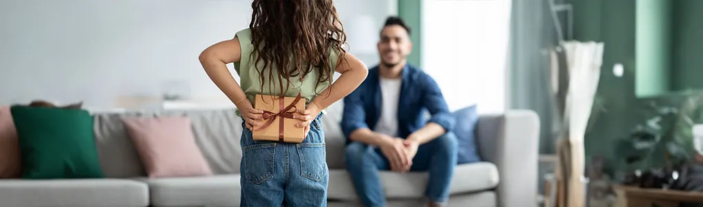 A child hides a wrapped gift with a ribbon behind their back from a seated adult in a denim shirt in a living room.
