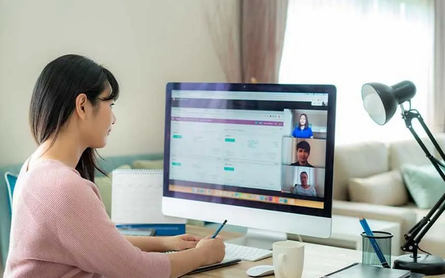 Een vrouw volgt een online vergadering op een groot bureauscherm, met meerdere videovensters geopend in haar lichte woonkamerkantoor.